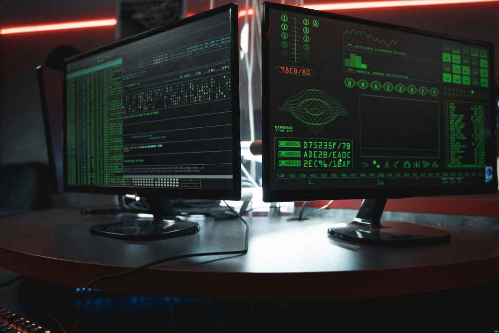 ¿Cómo proteger tu empresa de virus informáticos? Close-up of dual computer monitors with green coding interfaces in a dark room, highlighting cyber security themes.